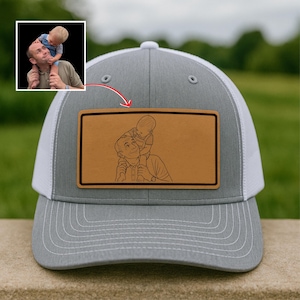Puede incluir: Gorra de camionero gris y blanca con un parche rectangular marrón. El parche presenta un dibujo lineal de una persona que lleva a un niño sobre sus hombros. La gorra tiene una visera curva y una parte trasera de malla.
