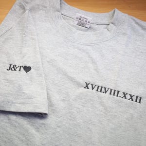 May include: A gray t-shirt with black embroidery. The left sleeve has "J&T" with a heart. The front of the shirt has "XVIII.VIII.XXII" embroidered in black.
