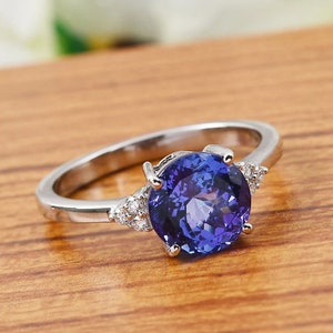 May include: A silver ring featuring a large, round, deep blue gemstone with a faceted surface. Small, clear accent stones are set on either side of the main stone. The ring is set against a warm, wooden surface.