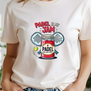 Op de afbeelding: Crèmekleurig T-shirt met een kleurrijke afbeelding. De afbeelding toont een pot jam met het woord "PADEL", geflankeerd door twee padelrackets en een bal. De tekst "PADEL IS MY JAM" staat boven de pot.