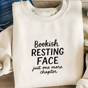 May include: A cream-coloured sweatshirt featuring the black text "Bookish Resting Face, just one more chapter". The folded sweatshirt displays the collar and sleeves. Denim shorts and a small plant are in the background.