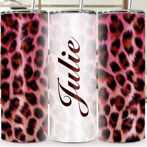 May include: Three stainless steel tumblers. Two have a pink and brown leopard print. The central tumbler has the name "Julie" in brown script on a white background. Each tumbler has a silver lid and a straw.