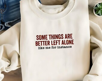 May include: Cream-colored sweatshirt with the text "SOME THINGS ARE BETTER LEFT ALONE like me for instance" in burgundy and black. The sweatshirt is folded, with a glimpse of denim shorts in the background.