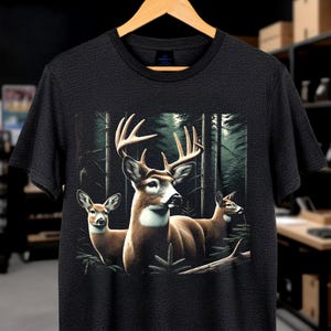 Puede incluir: Una camiseta gris carb&oacute;n de manga corta con un estampado gr&aacute;fico de tres ciervos en un bosque denso y oscuro. El ciervo central tiene astas grandes, mientras que los dos ciervos de los lados son m&aacute;s peque&ntilde;os. Incluye pinos y follaje.