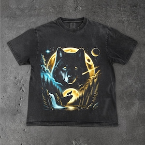 May include: Black t-shirt featuring a wolf design. The graphic includes a wolf's face in front of a full moon, with a smaller moon and a wolf silhouette on a cliff. The design incorporates a waterfall, trees, and stars.