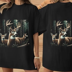 Puede incluir: Una camiseta negra de manga corta con un estampado gr&aacute;fico de dos ciervos marrones con astas en un bosque oscuro. Los ciervos est&aacute;n situados en el centro de la camiseta, rodeados de &aacute;rboles altos y follaje en un entorno boscoso profundo y sombr&iacute;o.