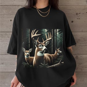 Puede incluir: Una camiseta negra de manga corta con un estampado gr&aacute;fico de tres ciervos en un bosque oscuro y denso. El ciervo central tiene astas grandes, mientras que los otros dos ciervos est&aacute;n situados a los lados. Tiene cuello redondo.