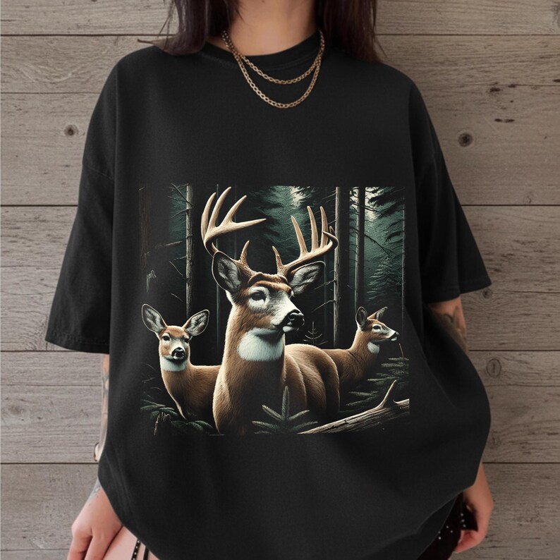 Puede incluir: Una camiseta negra de manga corta con un estampado gr&aacute;fico de tres ciervos en un bosque oscuro y denso. El ciervo central tiene astas grandes, mientras que los otros dos ciervos est&aacute;n situados a los lados. Tiene cuello redondo.