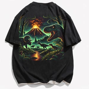 Può includere: Una t-shirt nera a maniche corte con una vivace stampa grafica di un paesaggio preistorico. La scena raffigura un grande vulcano in eruzione, una lussureggiante vegetazione giungla, un fiume che scorre e vari dinosauri, tra cui un T-Rex.