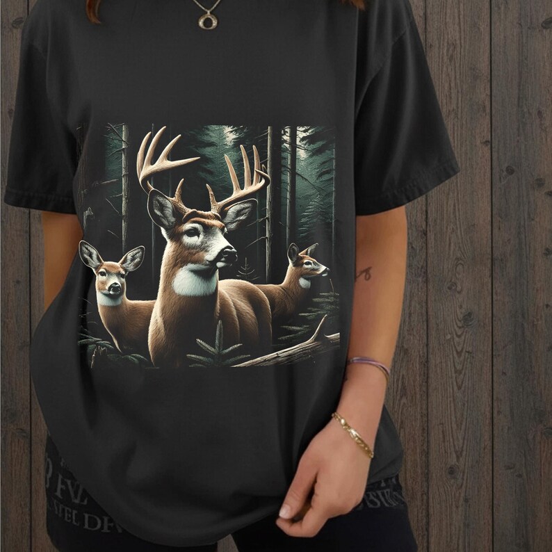 Puede incluir: Una camiseta negra de manga corta con un estampado gr&aacute;fico de tres ciervos en un entorno forestal. El ciervo central tiene astas grandes, mientras que los otros dos est&aacute;n situados a los lados. El fondo del bosque incluye &aacute;rboles altos.