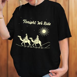 Könnte beinhalten: Schwarzes T-Shirt mit cremefarbener Grafik. Die Grafik zeigt zwei Personen auf Kamelen unter Sonne und Sternen, mit dem Text "Tonight We Ride". Das Design ist einfach und stilisiert.