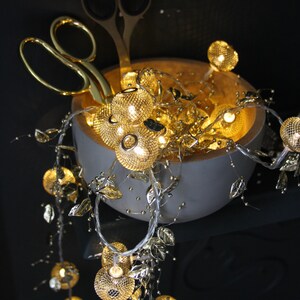 May include: A white ceramic bowl filled with gold fairy lights and gold leaf accents. The bowl is sitting on a dark surface and the lights are cascading down the side of the bowl.