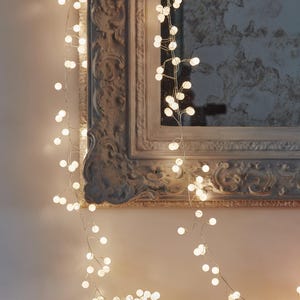 May include: A string of warm white, spherical fairy lights hangs near a decorative mirror. The lights are arranged to create a soft, ambient glow. The mirror has an ornate, vintage-style frame. Ideal for home decoration.