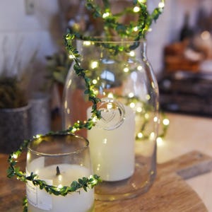May include: A string of fairy lights wrapped around two glass jars with white candles inside. The lights are surrounded by green leaves, creating a festive and cosy atmosphere.