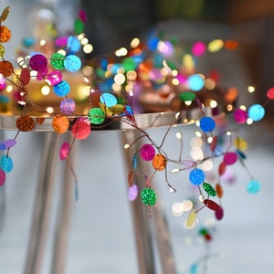 May include: A decorative string of colourful, sequined circles with small lights. The sequins are in shades of blue, green, orange, pink, and purple. The lights are small and warm, creating a festive display. The string is on a silver stand.