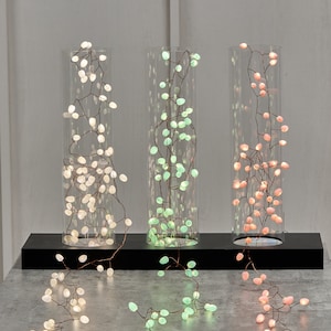 May include: Three strands of fairy lights with small, round bulbs in white, green, and pink. The lights are strung on a copper wire and are displayed in glass cylinders on a black surface.