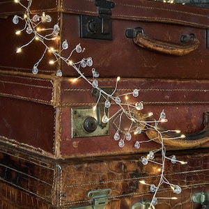 May include: Three vintage brown leather suitcases with a string of fairy lights wrapped around them. The lights have small clear glass bulbs.