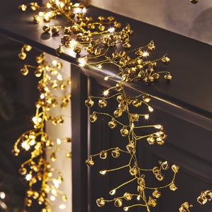 May include: A gold garland with small bells and warm white lights. The garland is draped over a dark surface, creating a festive and elegant look.