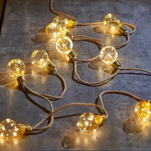 May include: A string of warm white LED lights with a rustic rope design. The lights are shaped like traditional incandescent bulbs and are strung together with a natural jute rope.