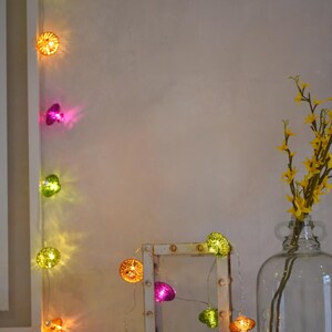 May include: A string of colourful, glowing decorative lights in shades of green, orange, and pink. The lights are shaped like small, textured orbs. A rustic, white-framed picture holder and a glass vase with yellow flowers are also in the image.