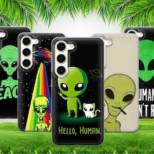 Puede incluir: Una colección de fundas para teléfonos con diseños de extraterrestres. Las fundas muestran extraterrestres verdes con varias expresiones, acompañados de frases como "PEACE" y "HELLO, HUMAN". Los fondos son negros, con un arcoíris y un OVNI en algunos.