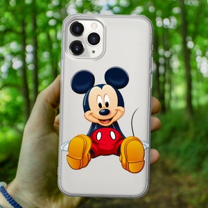 May include: A clear phone case with a Mickey Mouse design. Mickey Mouse is sitting down with his arms and legs spread out. He is wearing red shorts with two black circles and yellow shoes. Mickey Mouse is smiling and has black eyes, a black nose, and a black outline.