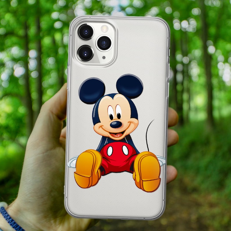 May include: A clear phone case with a Mickey Mouse design. Mickey Mouse is sitting down with his arms and legs spread out. He is wearing red shorts with two black circles and yellow shoes. Mickey Mouse is smiling and has black eyes, a black nose, and a black outline.