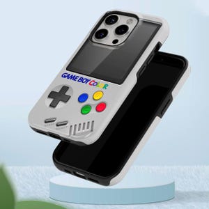 Może przedstawiać: Biało-szare etui na telefon zaprojektowane tak, aby przypominało Game Boy Color. Etui posiada pad kierunkowy, kolorowe przyciski oraz napis "GAME BOY COLOR". Etui na telefon jest pokazane z czarnym telefonem w środku.