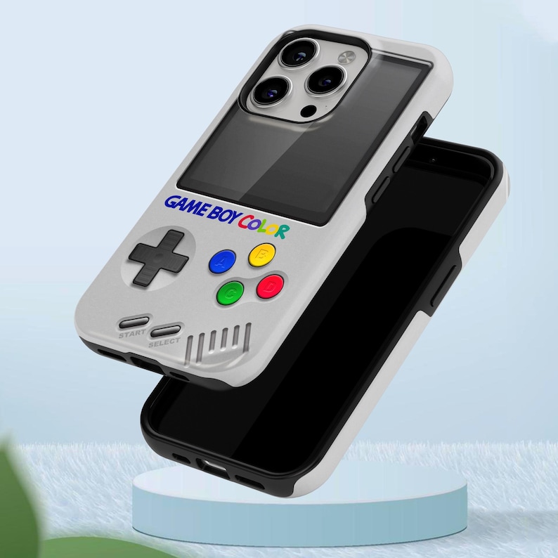 Może przedstawiać: Biało-szare etui na telefon zaprojektowane tak, aby przypominało Game Boy Color. Etui posiada pad kierunkowy, kolorowe przyciski oraz napis "GAME BOY COLOR". Etui na telefon jest pokazane z czarnym telefonem w środku.