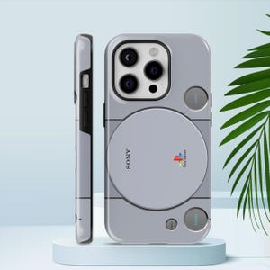 May include: A light grey phone case with a design inspired by the original PlayStation console. The case has the PlayStation logo and the word "SONY" printed on it. The case also has buttons labelled "OPEN" and "POWER".