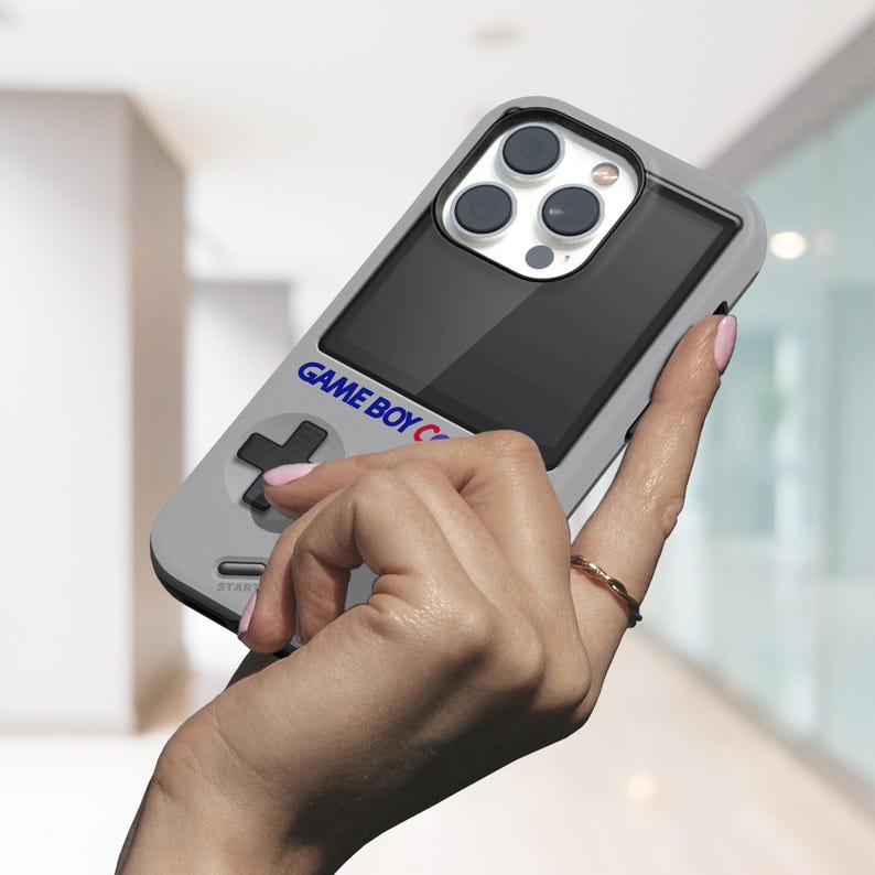 Może przedstawiać: Szara obudowa telefonu zaprojektowana na wz&oacute;r Game Boya. Obudowa posiada czarny ekran, krzyżak oraz niebieski napis "GAME BOY C". Obiektywy aparatu telefonu są widoczne na g&oacute;rze.