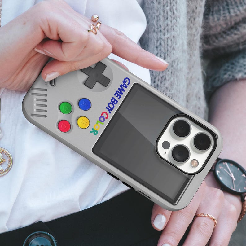 Może przedstawiać: Szare etui na telefon zaprojektowane tak, aby przypominało Game Boy Color. Etui posiada krzyżak, kolorowe przyciski oraz napis "GAME BOY COLOR" w tęczowych kolorach. Widoczne są soczewki aparatu telefonu.