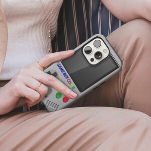 Może przedstawiać: Szara obudowa telefonu zaprojektowana tak, aby przypominała Game Boy Color. Obudowa posiada ekran, panel kierunkowy i kolorowe przyciski. Tekst "GAME BOY COLOR" jest wydrukowany z boku. Obudowa telefonu jest trzymana przez osobę.