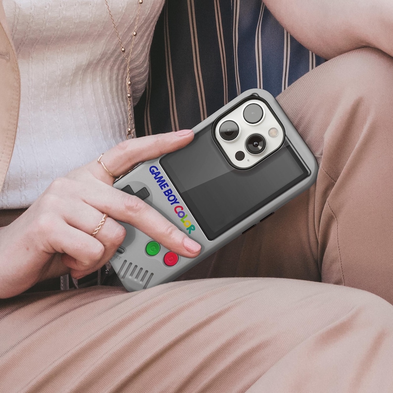 Może przedstawiać: Szara obudowa telefonu zaprojektowana tak, aby przypominała Game Boy Color. Obudowa posiada ekran, panel kierunkowy i kolorowe przyciski. Tekst "GAME BOY COLOR" jest wydrukowany z boku. Obudowa telefonu jest trzymana przez osobę.