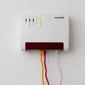 May include: A white FRITZ!Box router mounted on a wall, featuring a red ventilation grille at the bottom. The device has five vertical status indicator lights, with the two rightmost lights glowing green. Three ethernet cables in yellow, red, and white are plugged into the bottom ports.