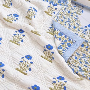 May include: A white quilted bedspread featuring a repeating pattern of blue flowers with green stems. The fabric has a diamond-stitched texture and a folded corner revealing a blue and white striped border trim along the edge of the textile.