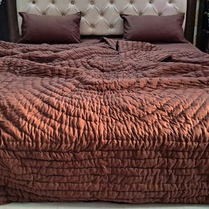 Può includere: Un letto con una trapunta marrone trapuntata e federe abbinate. La testiera è rivestita in un tessuto beige chiaro con un design a bottoni. Il letto si trova in una stanza ben illuminata.