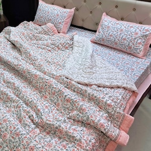 May include: A bedding set featuring a quilt and two matching pillowcases with a white base, decorated with a repeating pattern of small orange flowers and green leaves. The edges of the quilt and pillowcases are finished with a pink striped border.