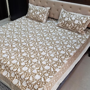May include: A white and gold floral patterned bedspread with a white and gold patterned pillowcase on a bed with a brown headboard.
