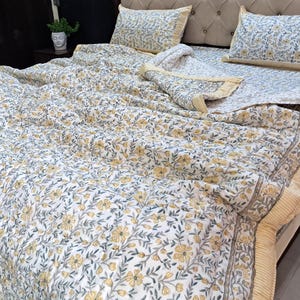Könnte beinhalten: Ein Bett ist mit einer floral gemusterten Steppdecke, zwei passenden Kissenbezügen und einem Spannbetttuch bezogen. Die Bettwäsche hat einen weißen Hintergrund mit gelben Blumen und grauen Blättern. Die Ränder der Steppdecke und Kissenbezüge haben einen hellgelben Rand.