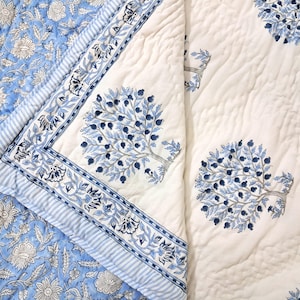 May include: A quilted cotton blanket with a blue and white floral and tree design. The blanket features a white background with blue tree and berry motifs, and a blue floral pattern on the reverse side.