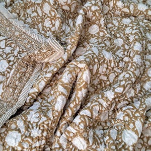 May include: A quilted blanket with a floral pattern in white and tan. The blanket features a detailed border with a striped design. The fabric is soft and appears to be cotton. The overall design is a traditional style, suitable for a bed or sofa.
