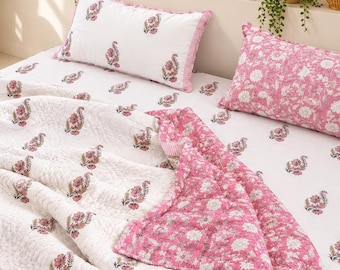May include: A white cotton quilt and matching pillowcases featuring a repeating pink and green floral paisley pattern. The quilt is folded back to reveal a solid pink underside with a white floral print, and the bedding set is laid out flat.