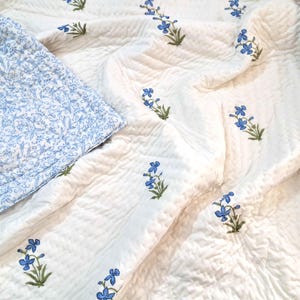 May include: A white quilted blanket featuring a repeating pattern of small blue flowers with green stems. One corner is folded over to reveal a contrasting underside with a dense, intricate light blue and white floral vine print pattern.