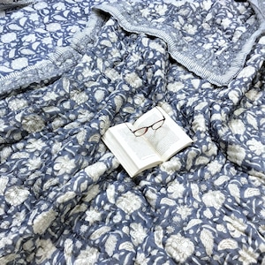 May include: A blue and white floral patterned quilt with an open book and eyeglasses resting on top. The quilt features a repeating floral design and a striped border. The book has white pages and the glasses have a brown frame.