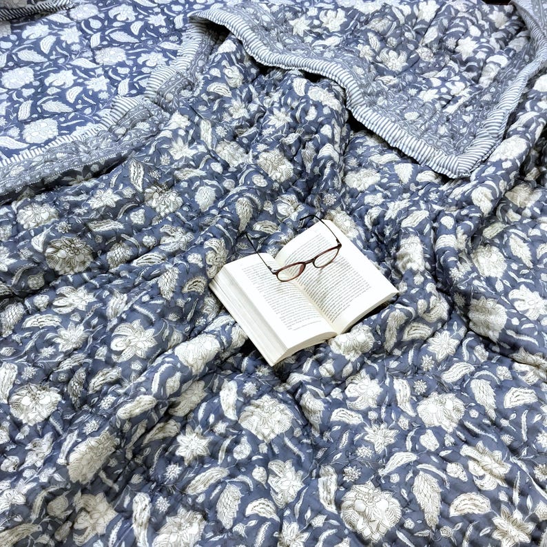 May include: A blue and white floral patterned quilt with an open book and eyeglasses resting on top. The quilt features a repeating floral design and a striped border. The book has white pages and the glasses have a brown frame.