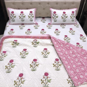 May include: A white bedding set featuring two rectangular pillows and a quilted coverlet, all decorated with a repeating pattern of pink flowers and green stems. The coverlet has a diamond-stitched texture and a solid pink floral reverse side.