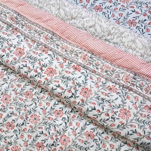 Op de afbeelding: Een gewatteerd textiel met een witte achtergrond en een herhalend patroon van kleine rode bloemen en grijze bladranken. De stof bevat een opvallende randsectie met dunne rode strepen en een bloemmotief, wat een gelaagd visueel effect creëert.