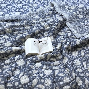 Könnte beinhalten: Eine blau-weiß gemusterte Steppdecke mit passendem Rand. Ein offenes Buch mit Brille liegt mittig auf der Steppdecke. Die Steppdecke ist aus weichem Material und für ein Bett geeignet.