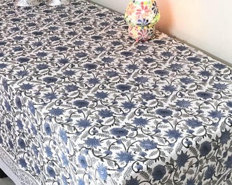 Puede incluir: Un mantel rectangular blanco con un estampado floral azul. El diseño presenta flores azules estilizadas y hojas grises sobre un fondo blanco. El mantel está sobre una mesa con una pequeña lámpara y cuencos de vidrio decorativos.